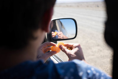 Cropped image of man peeling orange while sitting in car by reflection on side-view mirror