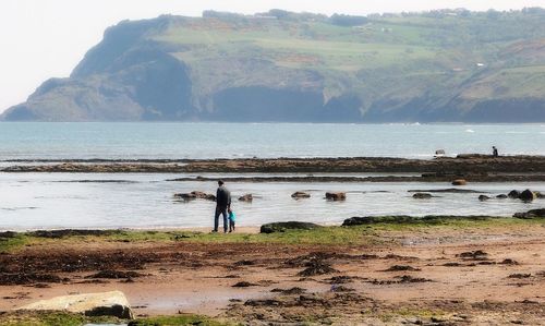 Father and son standing on rock by sea against mountain