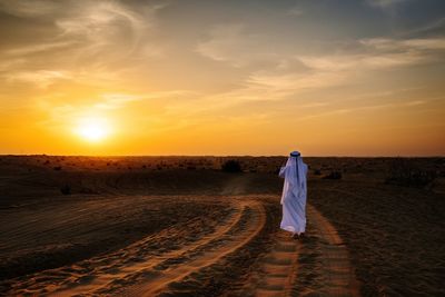 Rear view of man wearing white traditional clothing walking in desert during sunset