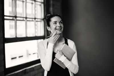 Smiling woman holding book while standing against wall