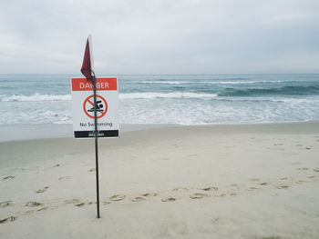 No swimming sign on sand at beach against cloudy sky