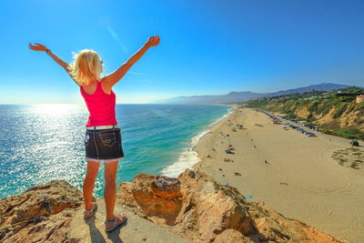 Rear view of mature woman with arms raised standing on cliff by sea against blue sky during sunny day