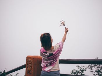 Rear view of woman standing by railing holding plant against sky