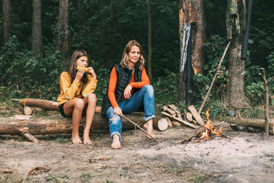 A mother with her teenage daughter is sitting on a tree near a campfire in the forest 