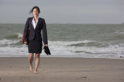 Business woman walking on empty beach holding her shoes