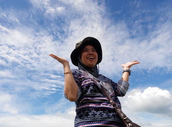 Low angle view of woman with hands raised laughing against cloudy sky