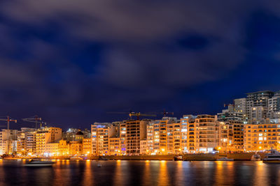Illuminated buildings by sea against sky at night