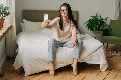 Young happy woman sitting on bed holding cellphone and communicating via video calls