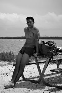 Full length portrait of shirtless man leaning on bench at beach against sky