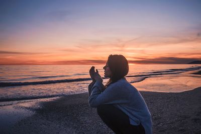 Side view of woman crouching at beach against sky during sunset
