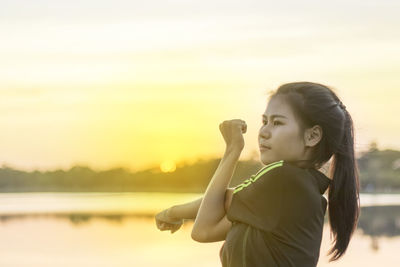 Side view of young woman exercising by sea against sky during sunset
