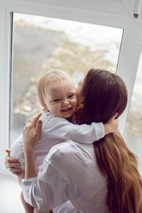 Mother in a white robe sits with a child a blonde daughter at a large window of the house person