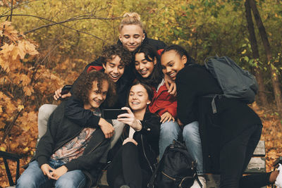 Teenage girl taking selfie with friends while sitting on bench against trees during autumn