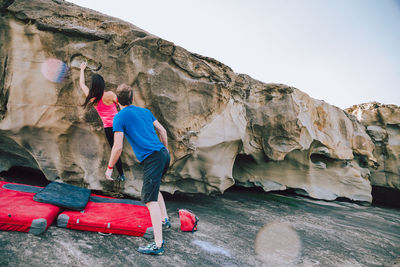 Man looking at woman climbing on rock at beach against sky