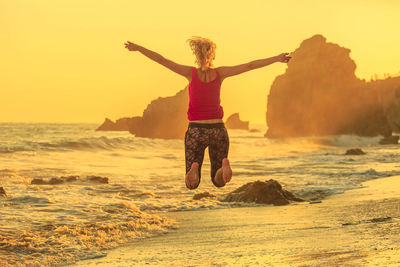 Rear view of mature woman jumping at beach against sky during sunset