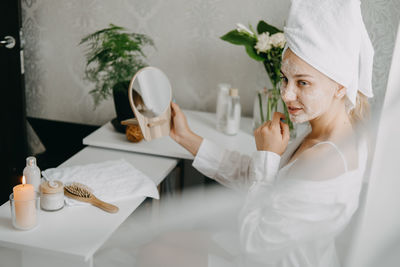Young woman in white towel chilling in bedroom and making clay facial mask near mirror.