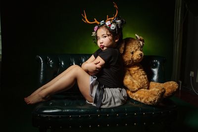 Full length portrait of angry girl with arms crossed sitting by teddy bear on sofa