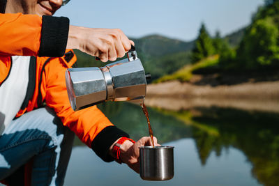 Midsection of man with coffee kettle pouring drink in cup standing by lake
