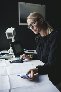 Businesswoman looking at blueprint while using calculator on desk