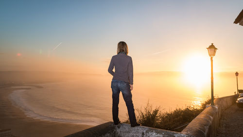 Rear view of woman standing on retaining wall overlooking sea against clear sky during sunset