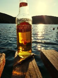 Glass of bottles on table by lake against sky