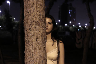 Portrait of woman standing behind tree trunk at night