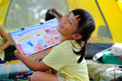 Girl painting on paper while sitting against tent