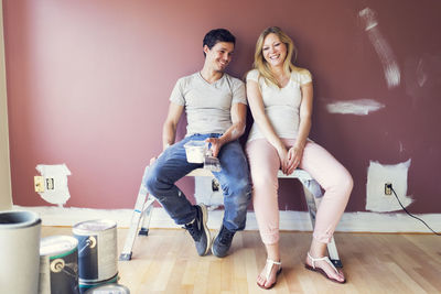 Happy couple sitting on stool against wall at home