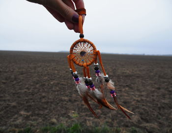 Cropped hand of woman holding dreamcatcher over field against sky