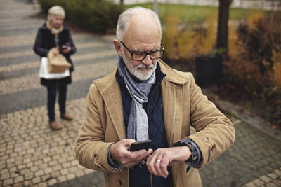 Senior man with smart phone checking the time while partner standing behind on footpath