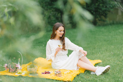 Beautiful young woman in a white dress drinking wine in the garden on a picnic