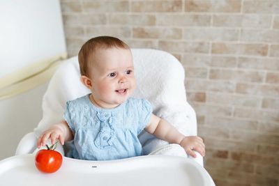 Cute baby girl with tomato sitting on high chair at home