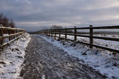 View along snowy footpath with fence in winter against sky 