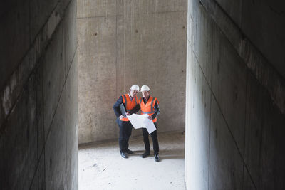 Two men with plan wearing safety vests talking in building under construction