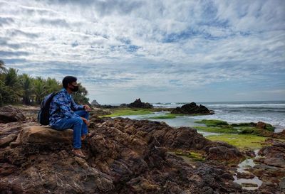 Side view of man sitting on rock by sea against sky