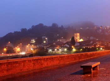 Illuminated street by buildings against sky at night