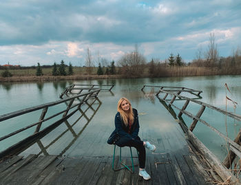 Young woman sitting on wood by lake against sky