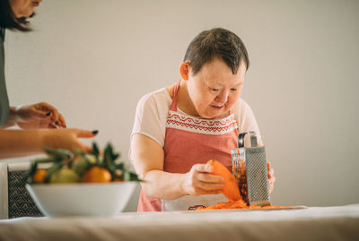 Elderly woman with down syndrome gratefully learns to grate carrots with the guidance of a teacher