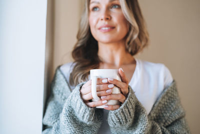 Young woman with blonde long curly hair in cozy grey sweater with cup of tea in hands at home