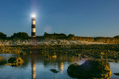 Illuminated lighthouse by lake and buildings against sky