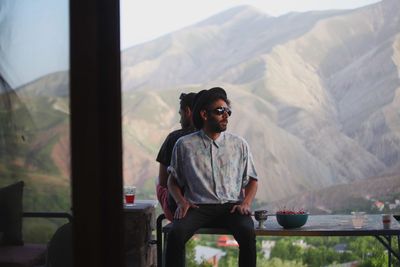 Male friends sitting back to back on bench against mountains