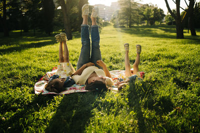 Happy young woman and adorable little sisters in similar dresses lying on blanket on green grass while spending summer day together in park and legs up