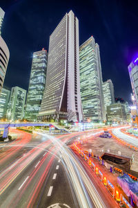 Light trails on city street by buildings against sky at night