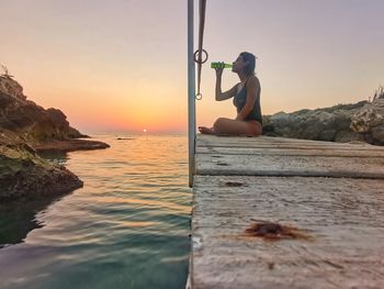 Woman sitting on rock by sea against sky during sunset