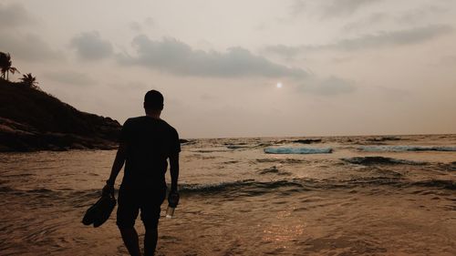 Rear view of man holding shoes while walking in sea against sky during sunset