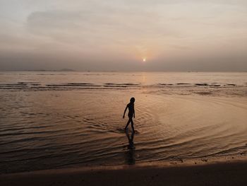 Silhouette boy standing at beach against sky during sunset