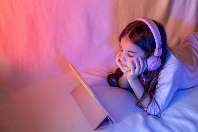 Little girl in pink headphones, lies on the bed , looks into a digital tablet
