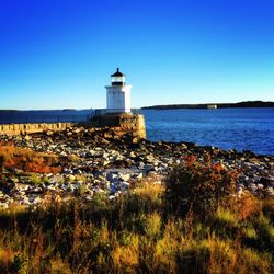 Lighthouse by sea against clear blue sky