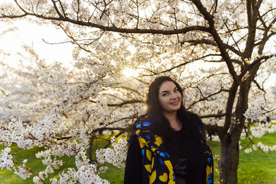 Young long-haired plus size woman spending time among cherry blossom trees in city park in spring