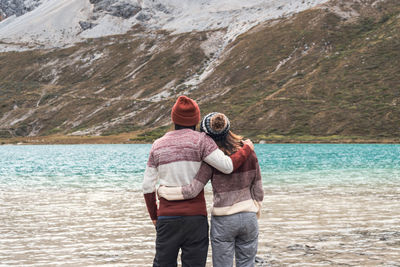 Rear view of couple standing by lake while looking at mountains during winter